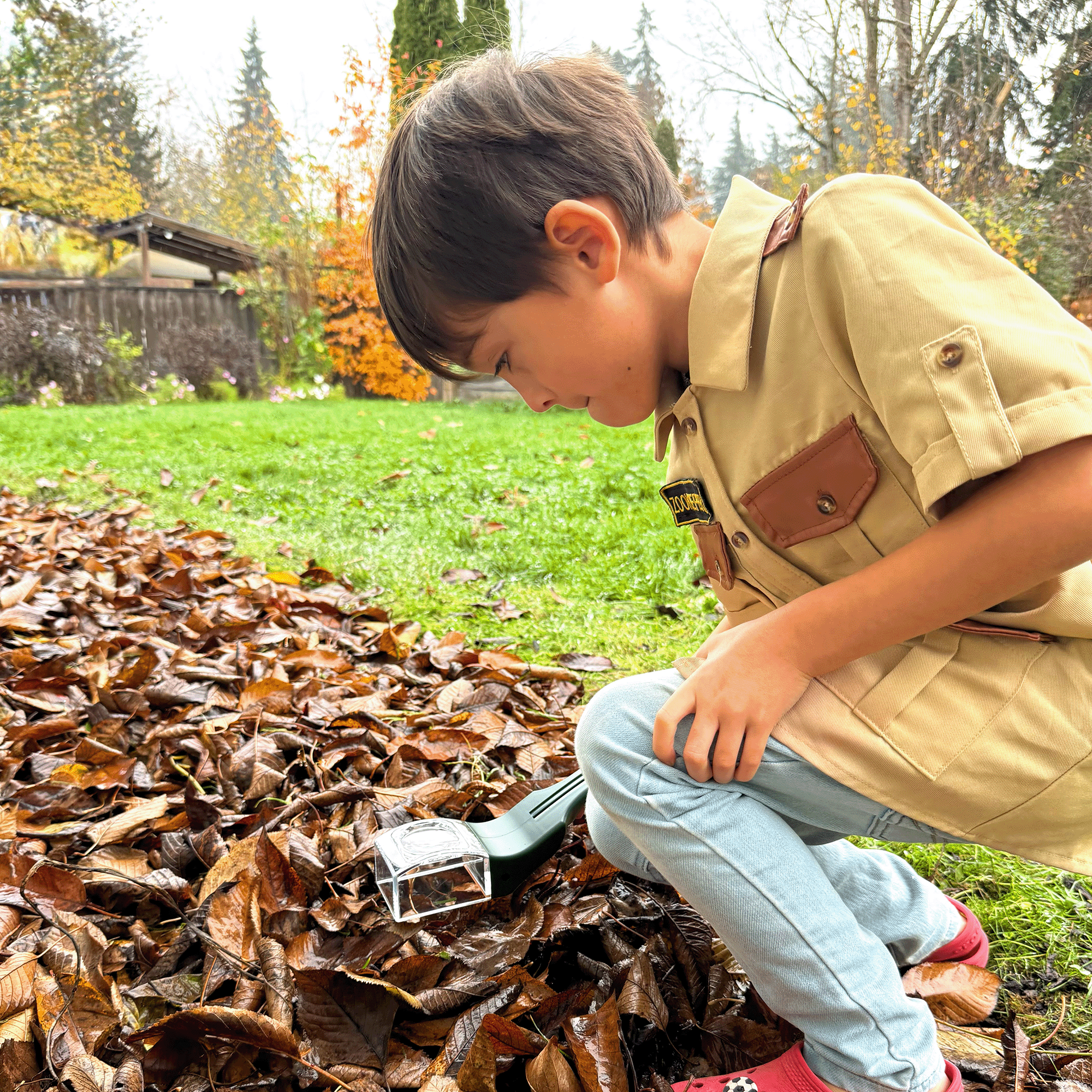 Child in a beige shirt and light blue pants crouching over a pile of leaves, holding a small tool.